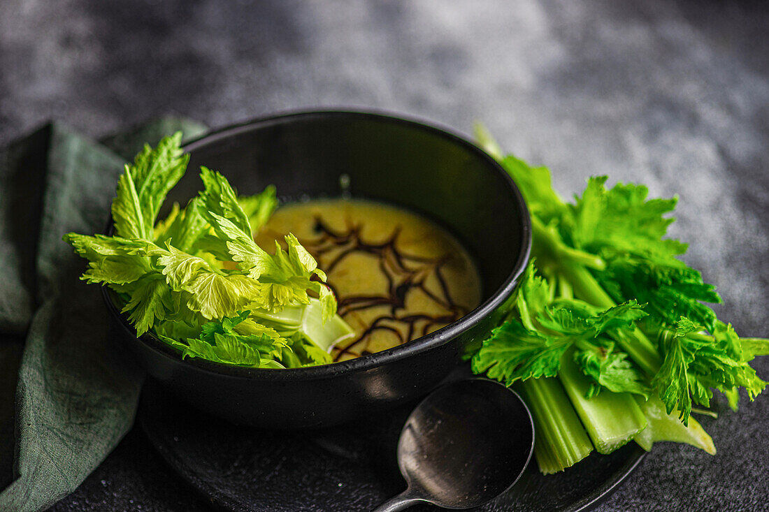 Front view of healthy cream of celery soup in a bowl with celery sticks served on a black plate with black spoon and napkin against a blurred dark background