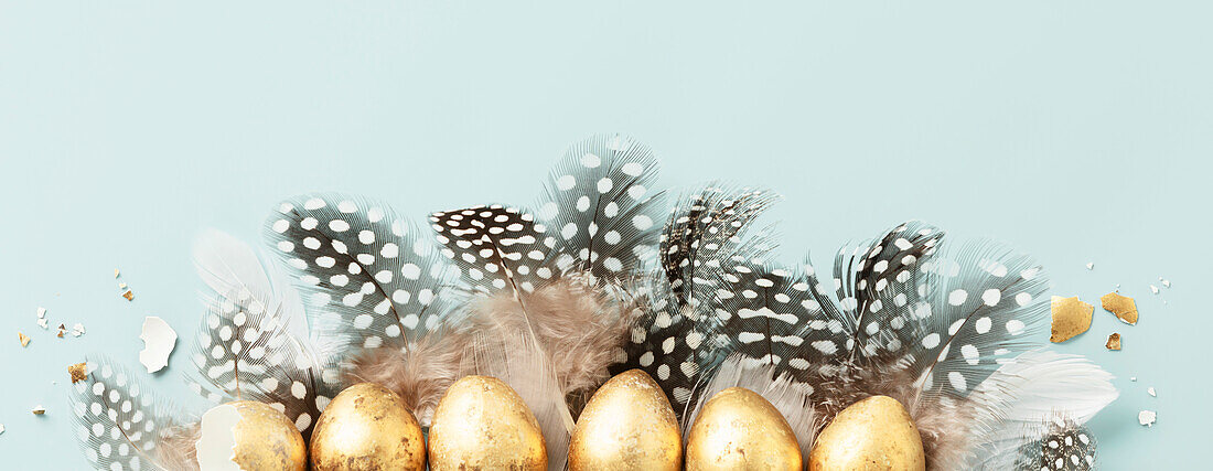Raw of golden quail eggs and quail feathers on a blue background. Beautiful modern minimalist Easter composition. Copy space, top view, flat lay