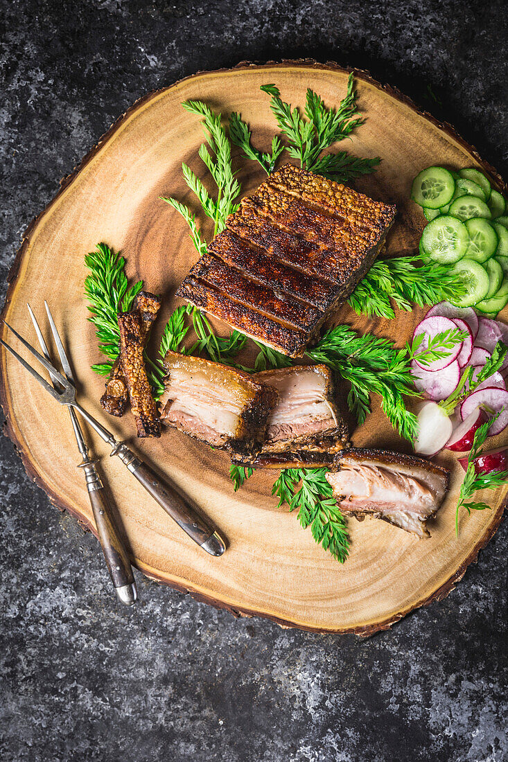 Sliced crispy pork belly on a wooden plate with forks, sliced gherkins and radishes