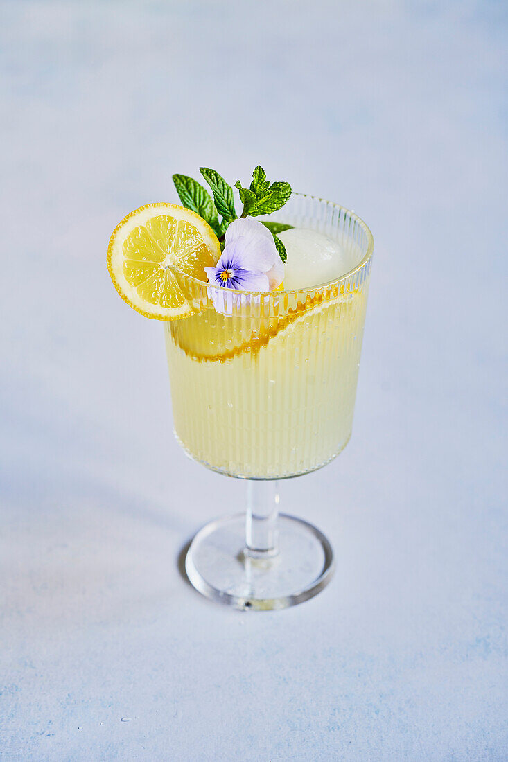Lemon and mint lemonade mocktail on a light blue background with lemon, mint and purple flower garnish