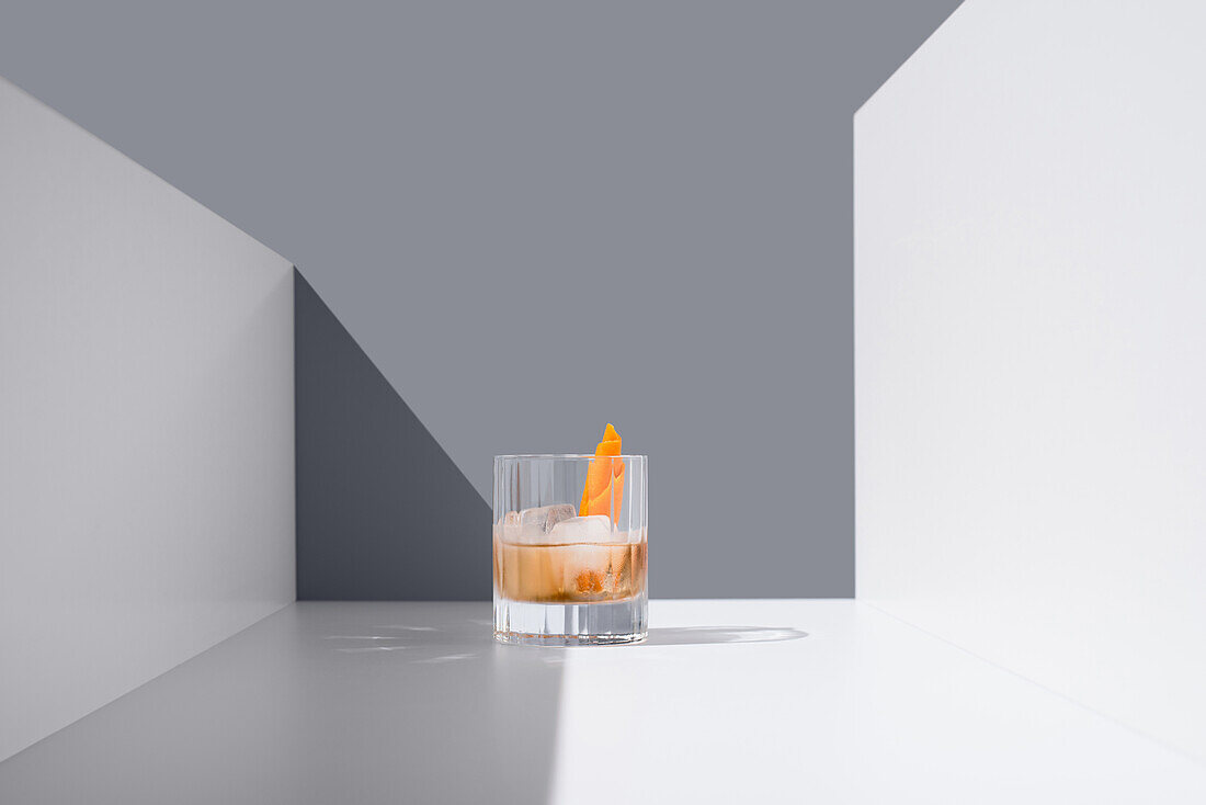 Crystal clear glass filled with fresh old fashioned cocktail garnished with orange and ice cubes placed on white surface between white walls against gray background
