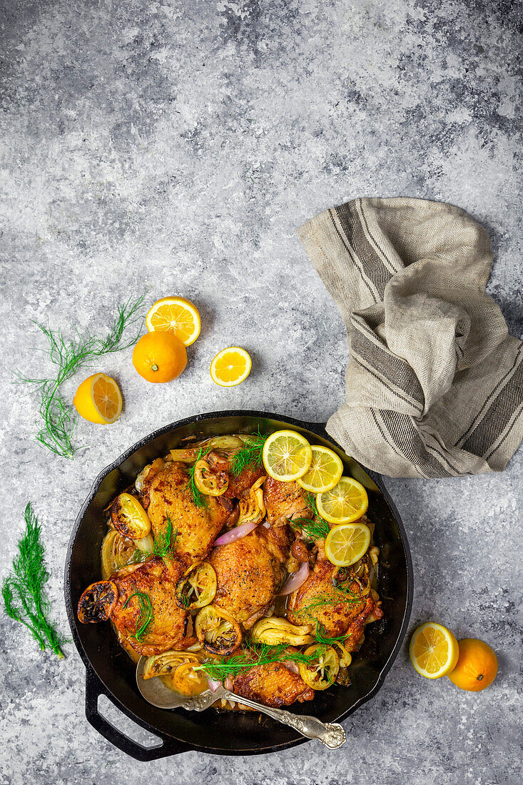 Chicken thighs in a cast-iron pan with fresh and roasted lemons, fennel and shallots on a grey background with lemon slices as garnish