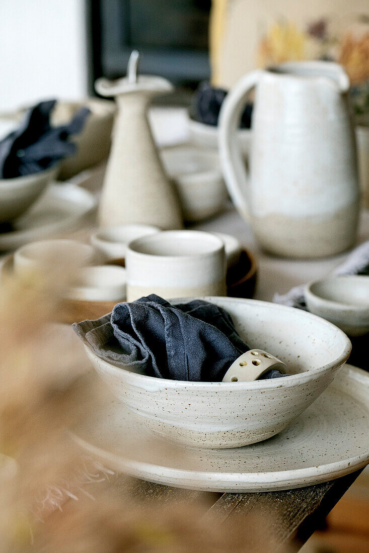 Rustikaler Tisch mit leerem, handgefertigtem Keramikgeschirr, weißen, rauen Schüsseln, Tellern, Tassen, Krug und Vase auf einem Leinentischtuch. Getrocknete Schilfblüten. Tageslicht Esszimmer