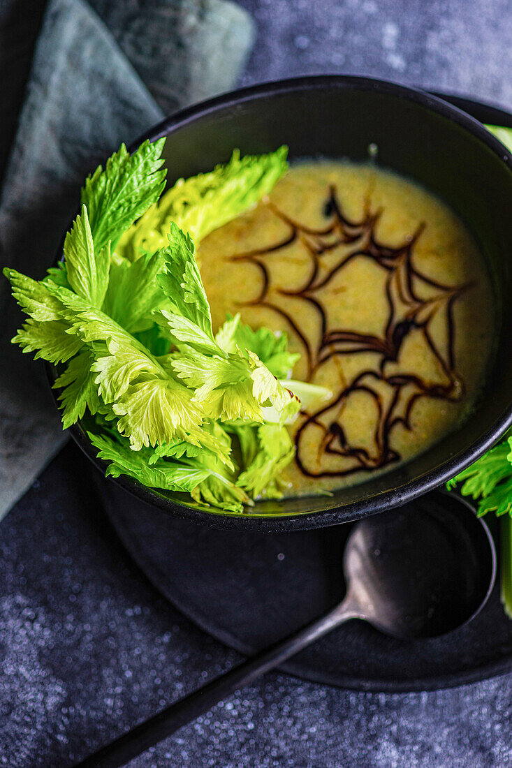 Healthy cream of celery soup in bowl with celery sticks on black plate with black spoon and napkin in front of blurred dark background