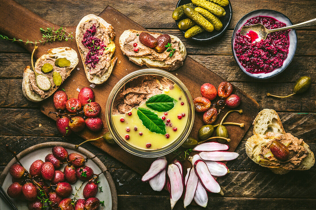 Pâté spread in a glass on a wooden board with roasted grapes, radishes, gherkins and baguette
