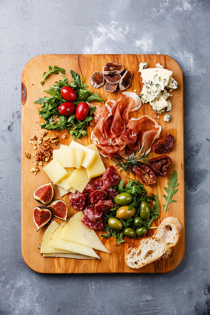 Italian snacks with ham, olives, cheese, sun-dried tomatoes, sausage and bread on a wooden cutting board against a concrete background