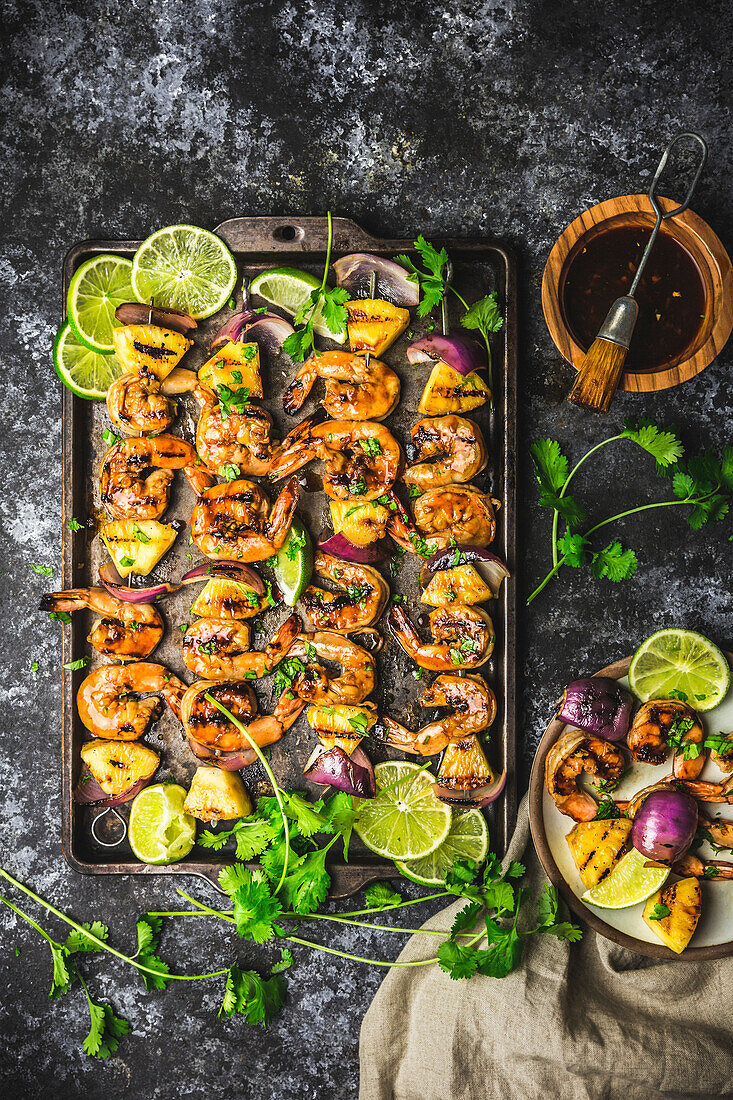 Skewers with grilled prawns, pineapple and red onions on a baking tray and plate with glaze in a bowl