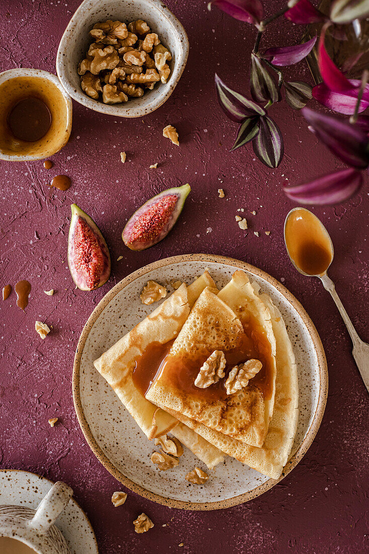 Crepes, folded on a handmade ceramic plate, decorated with figs, caramel and walnuts over a purple background