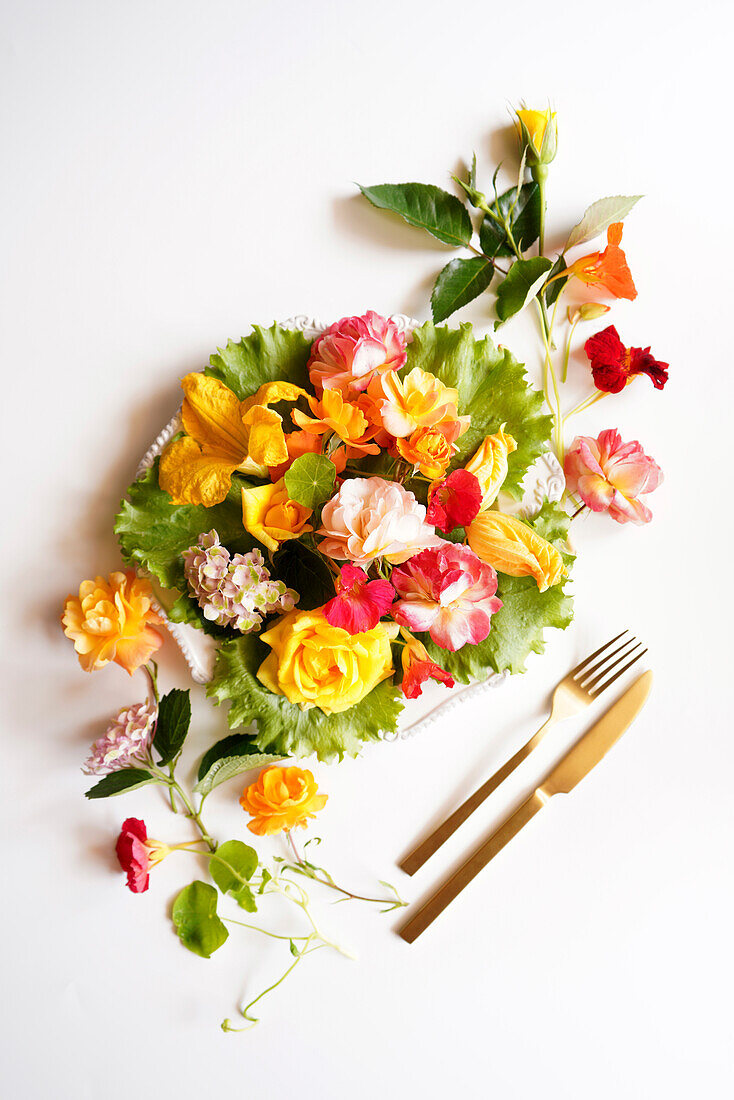 Essbare Blumen, darunter Rosen, Hortensien, Zucchini und Kapuzinerkresse, auf Teller mit Gabel und Messer, kreatives Konzept Flatlay