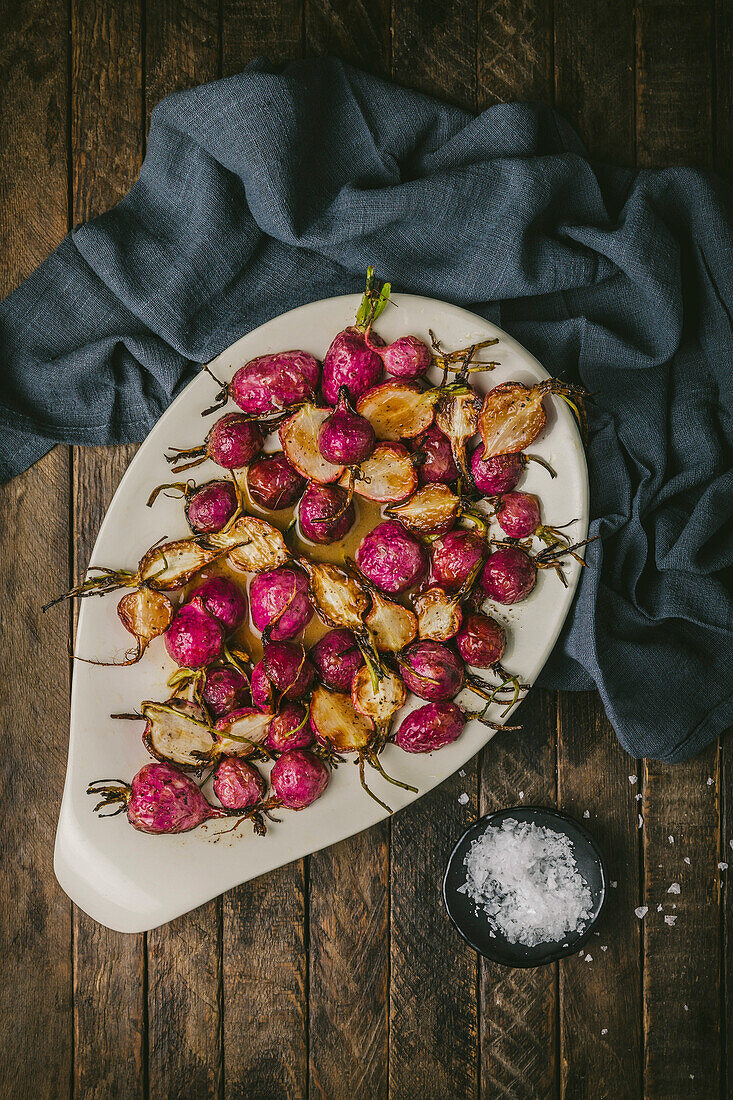 Oval platter with fried pink radishes in brown butter sauce on a wooden table with a napkin and sea salt