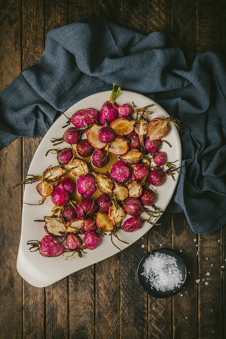 Ovale Platte mit gebratenen rosa Radieschen in brauner Buttersauce auf einem Holztisch mit einer Serviette und Meersalz