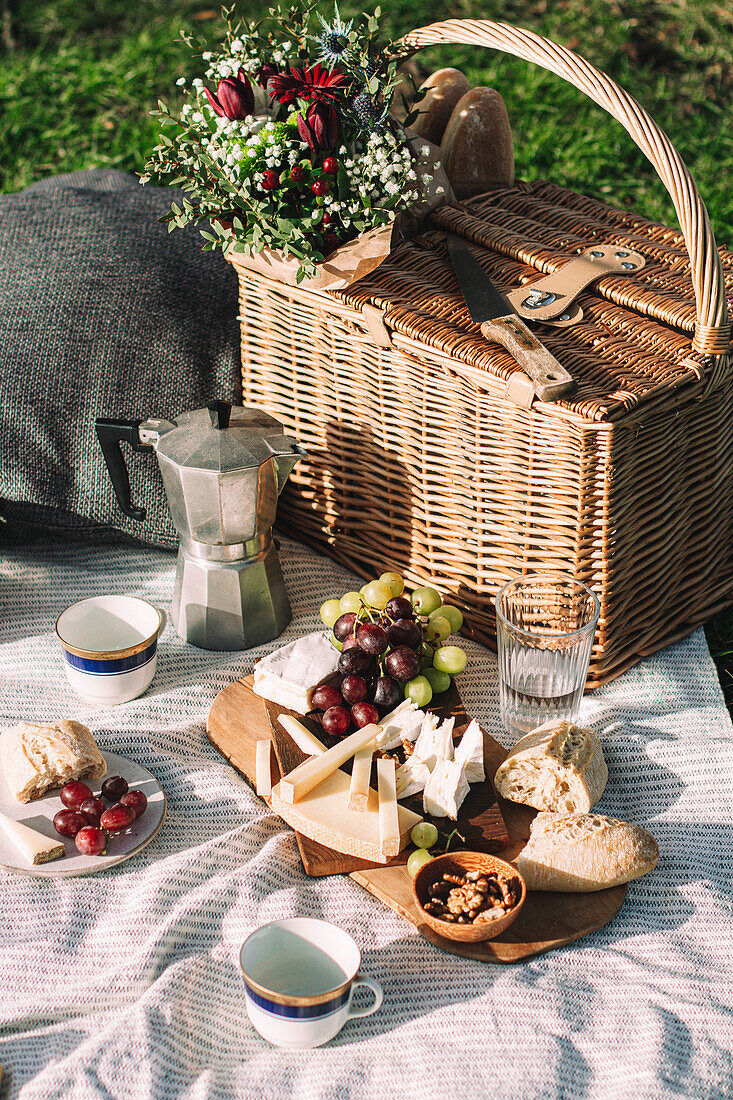 Picknickszene mit einem Korb, Kaffee und einer Tafel mit Weintrauben, Käse und einem Baguette