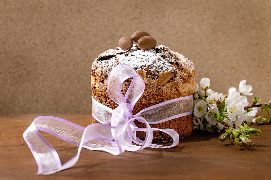 Homemade Italian traditional Easter panettone cake, decorated by chocolate eggs, pink ribbon and blossoming cherry tree flowers standing on wooden table. Traditional Easter European bake. Copy space
