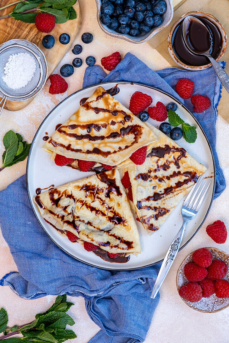 Crepes garniert mit Schokoladensauce, frischen Beeren und Minzblättern auf einem Teller, eine Gabel an der Seite