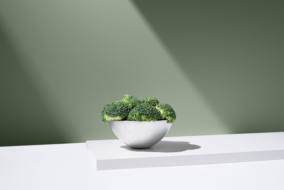 Frischer grüner Brokkoli wächst in einer weißen Schale auf einem schlichten Tisch vor grauem Hintergrund unter hellem Lichtstrahl