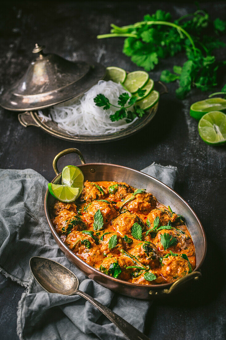 Meatballs in curry sauce in a serving bowl with noodles and lime wedges