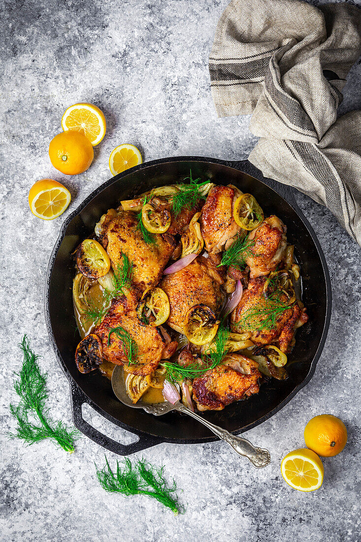 Hähnchenschenkel in gusseiserner Pfanne mit frischen und gerösteten Zitronen, Fenchel und Schalotten auf grauem Hintergrund