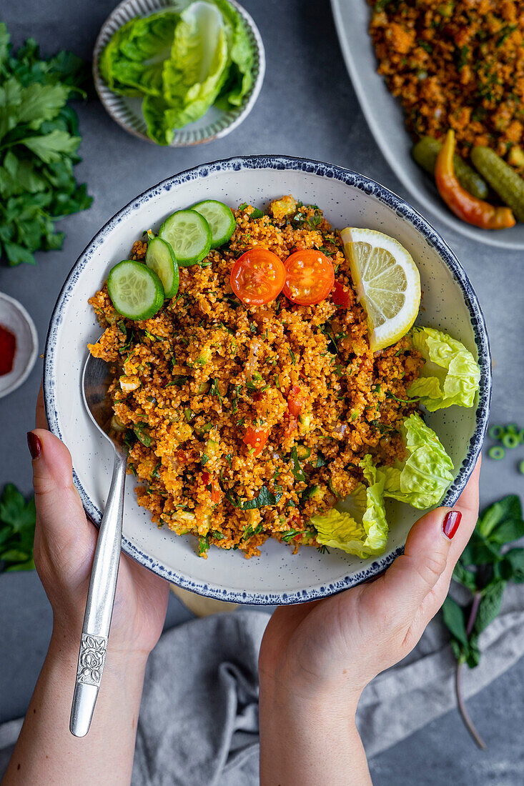 Woman hands holding Turkish bulgur salad garnished with sliced lemon, tomatoes and cucumbers in a white bowl.