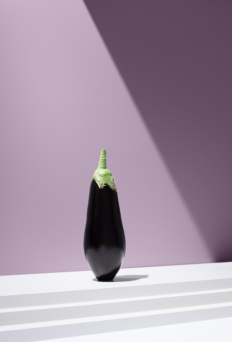 Vivid image of a black aubergine with a green cap on a white, tiered plate against a purple background under bright light