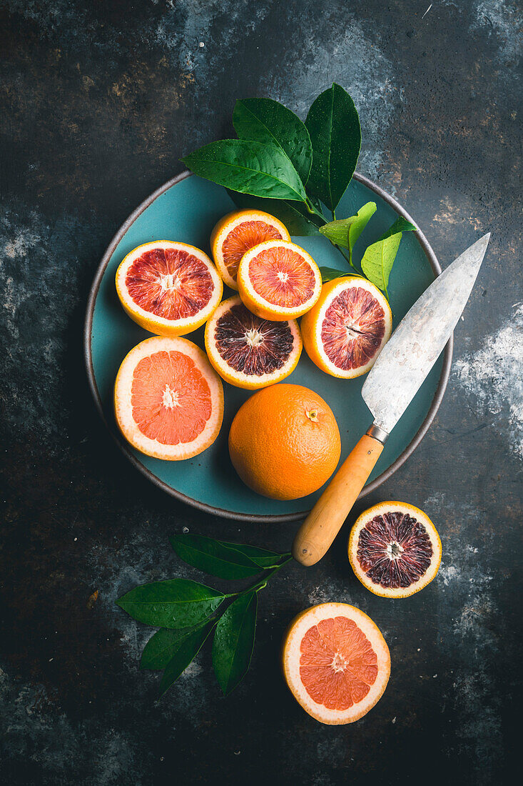 Blood oranges and pink grapefruit, halved and whole, with citrus leaves, in blue ceramic bowl and on table, with knife on dark background