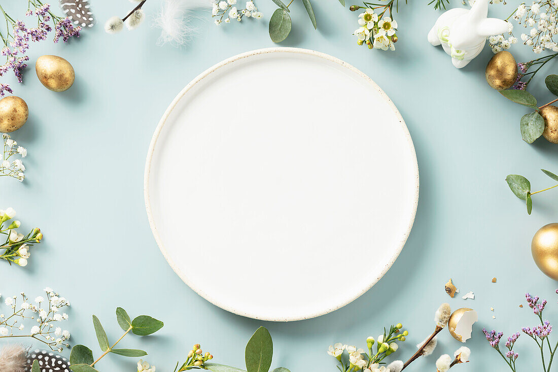 Easter table decoration. Happy Easter concept with white plate, golden Easter eggs, feathers and spring flowers on blue background. Flat lay top view copy space