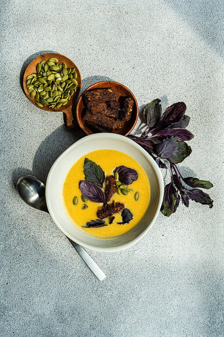 Top view of bowl with pumpkin cream soup with basil herb, rye bread and seeds on blurred grey background with leaves