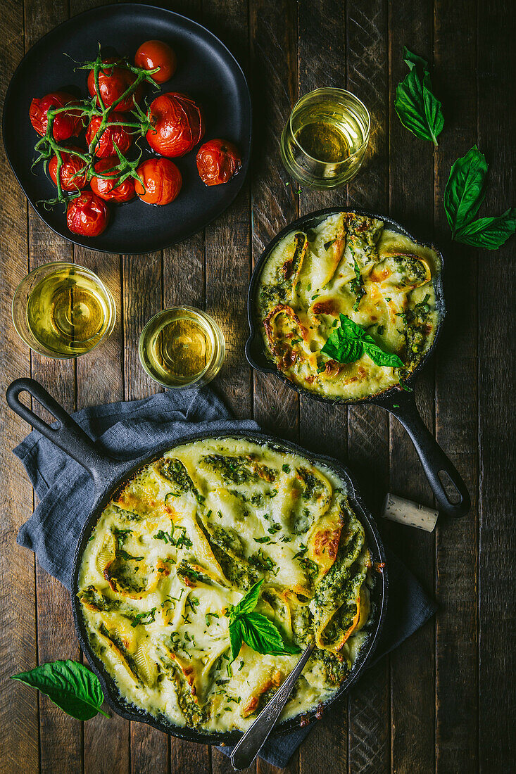 Small and large cast iron skillets with cheesy baked pasta, with roasted tomatoes, wine and basil garnish on wood table