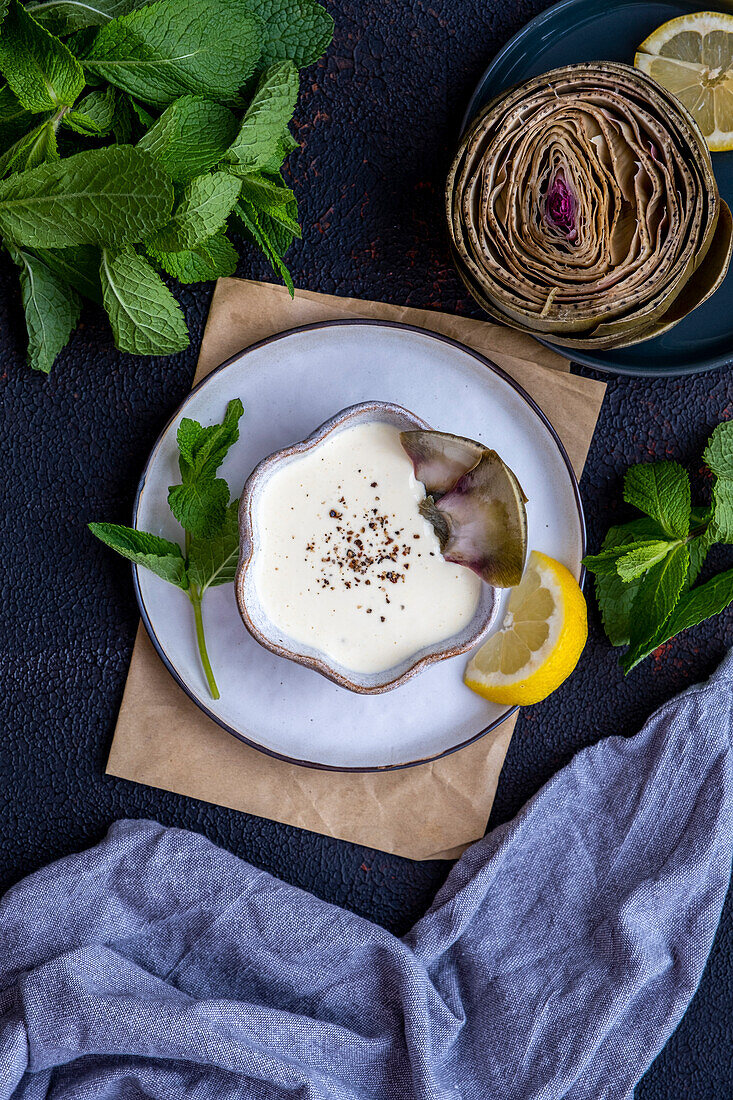 Eine Schale mit Artischocken-Dip-Sauce, garniert mit schwarzem Pfeffer und Artischockenblättern an der Seite, von oben fotografiert