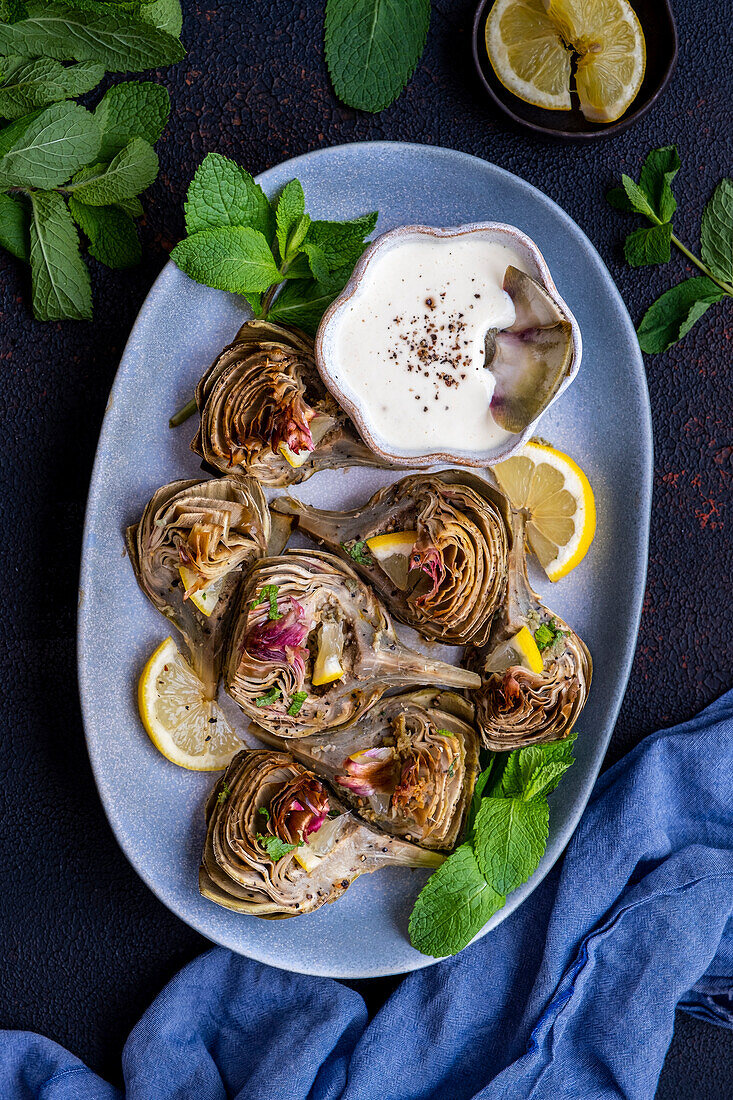 Roasted artichokes served on an oval plate with a bowl of dipping sauce on the side.