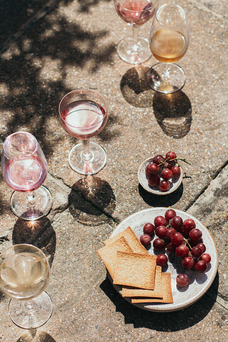 Gläser mit Rosé- und Weißwein, Weintrauben und Crackern, aufgenommen in hartem Licht auf Fliesen
