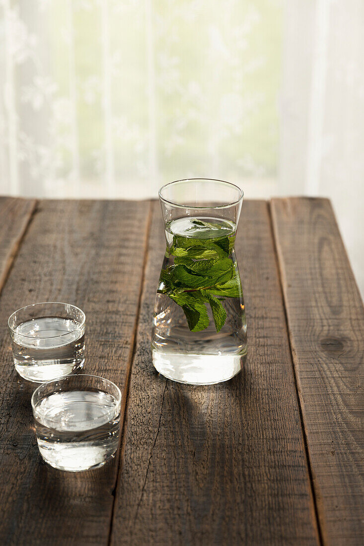 Frisches Wasser und Minzblätter in einer Karaffe und zwei mit Wasser gefüllten Bechern, auf einem Holztisch