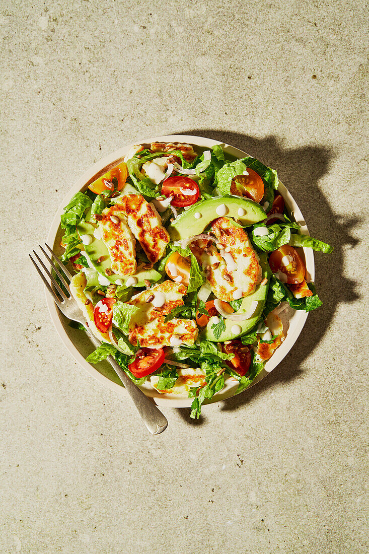 Halloumi Avocado Tomato Salad with sunflower seeds, yogurt dressing, herbs and water on a green background with shadow