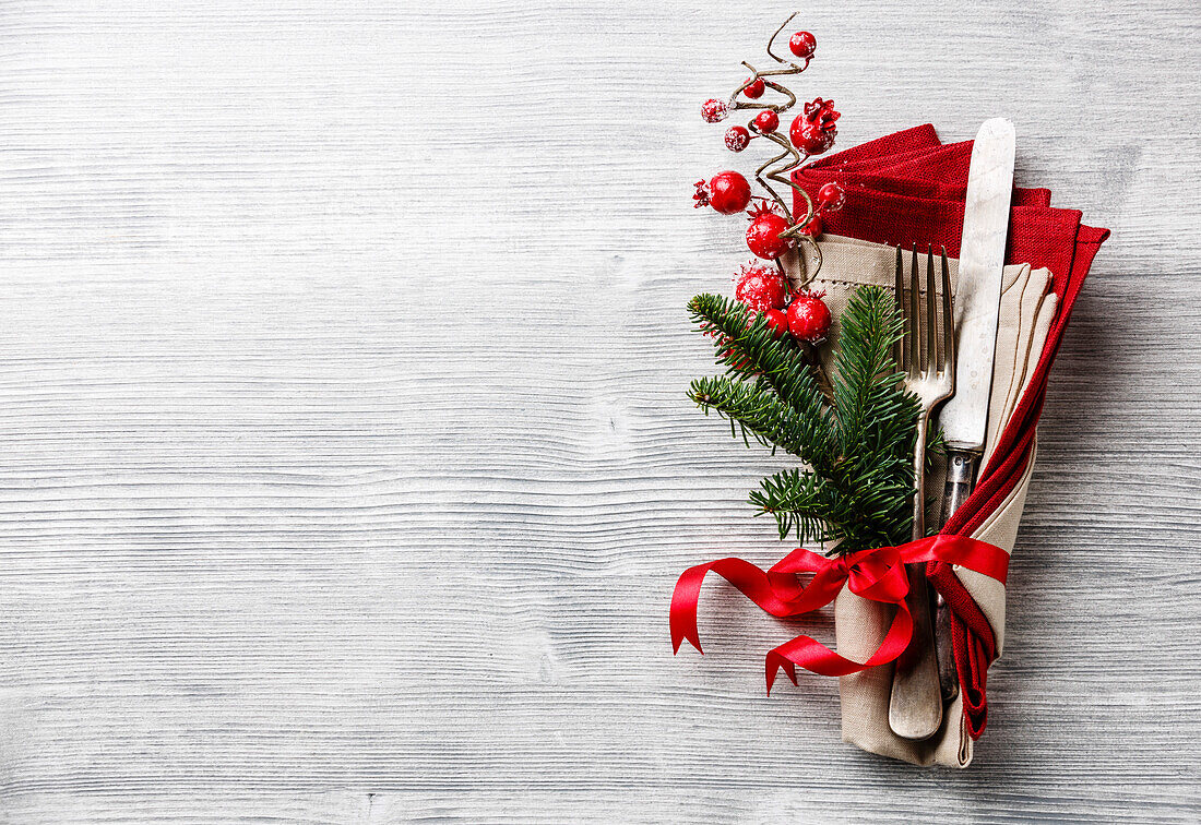Table fork and knife set with napkin, Christmas fir branch, red berries and ribbon on grey wooden background Copy Space