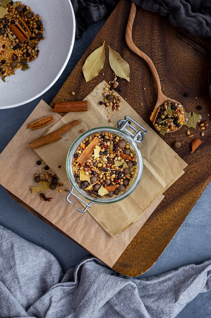 Mustard seeds, coriander seeds, cloves, cinnamon sticks, bay leaves and red pepper flakes mixed in a glass jar with a wooden spoon inside