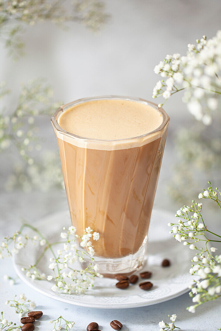 A vegan latte in a tall, heat-resistant glass surrounded by white flowers