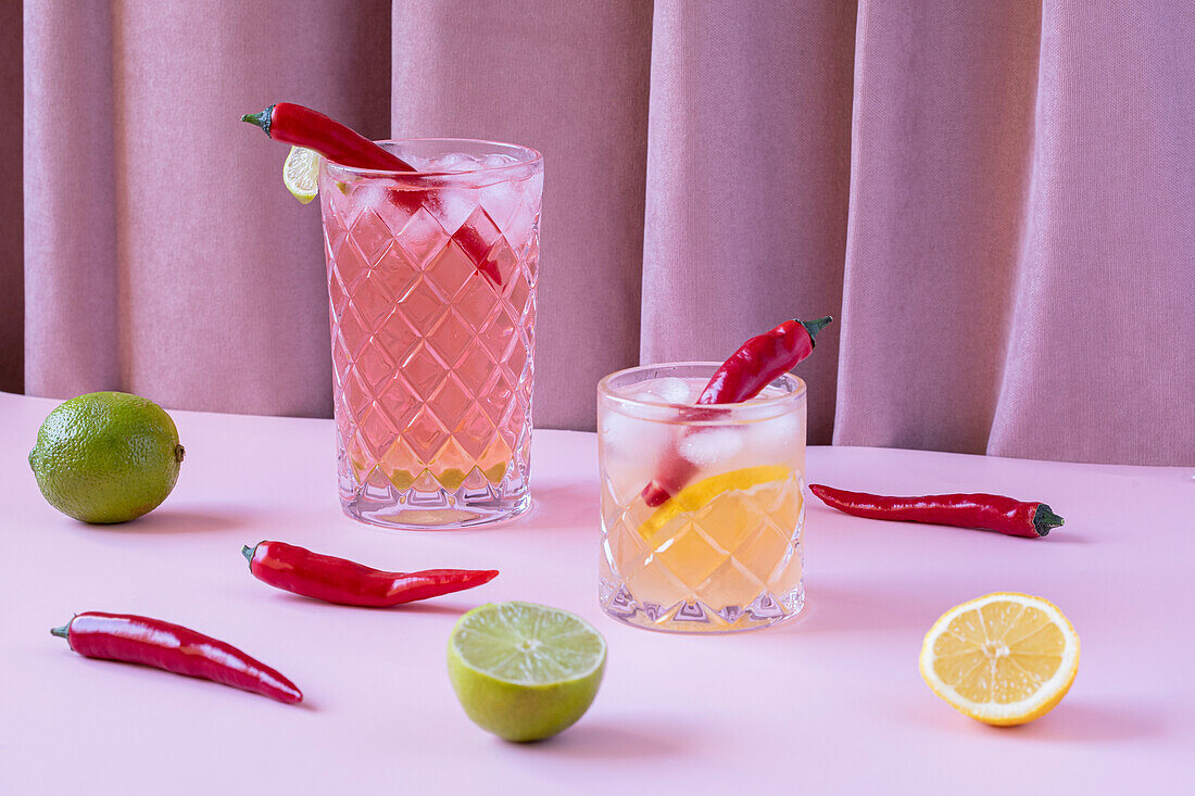 Transparent glasses filled with cold, refreshing cocktails, served with chilli peppers and ice cubes, on a table with lemons and limes