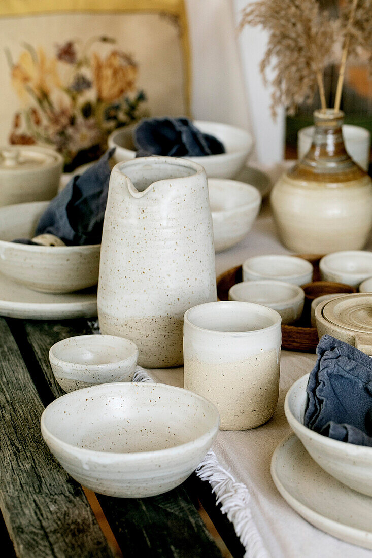 Rustic table with empty, handmade ceramic crockery, white, rough bowls, plates, cups, jug and vase on a linen tablecloth. Dried reed flowers. Daylight dining room