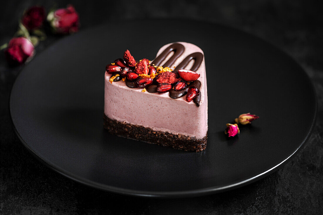 Heart-shaped cake decorated with pomegranate and strawberries. Dessert on a black plate with a dried rose