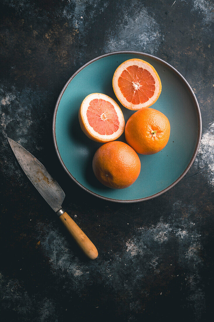 Rosa Grapefruit, eine halbiert und zwei ganz, in blauer Keramikschale mit Messer auf dunklem Hintergrund