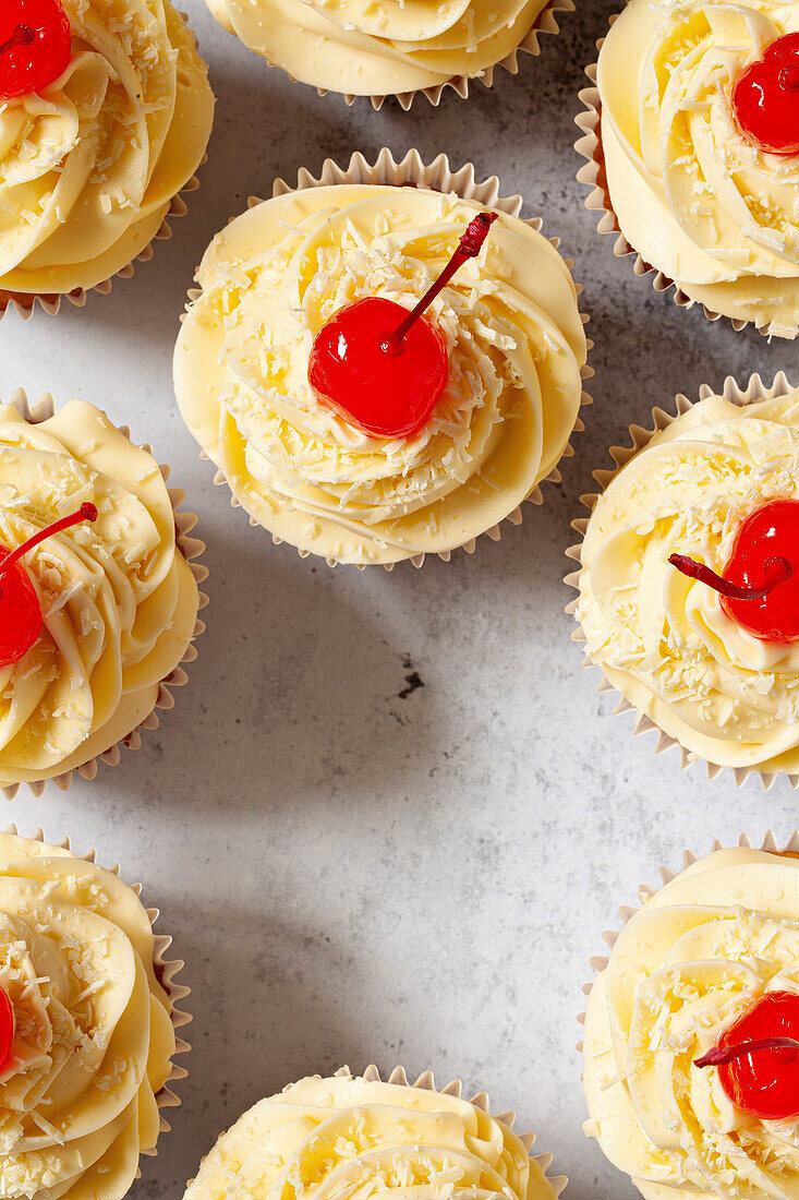 Cupcakes decorated with piped buttercream frosting, grated white chocolate and maraschino cherries with stems on.