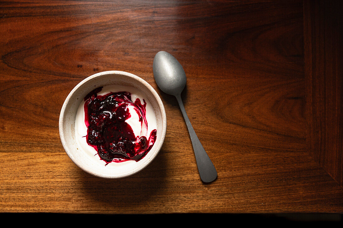 Bowl of yoghurt and blackberry compote on antique wooden table with silver spoon
