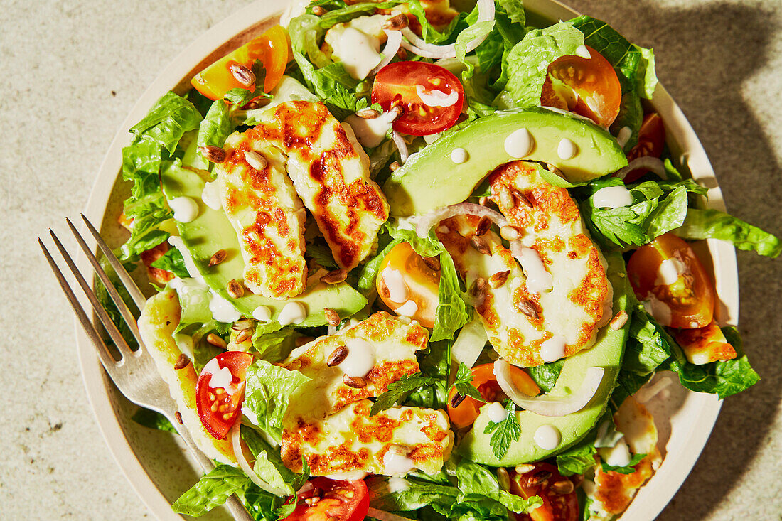 Halloumi avocado tomato salad with sunflower seeds, yoghurt dressing, herbs and water on a green background with shade