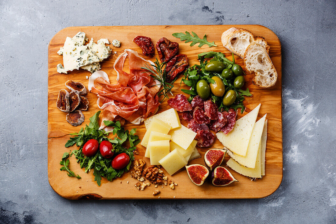 Italian snacks with ham, olives, cheese, sun-dried tomatoes, sausage and bread on a wooden chopping board in front of a concrete background