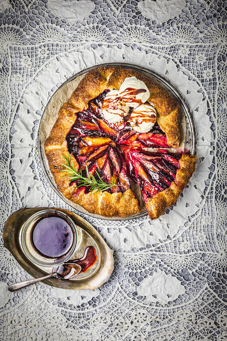 Rustic plum galette, sliced, with vanilla ice cream, red wine caramel sauce and a sprig of fresh rosemary on an intricate lace background