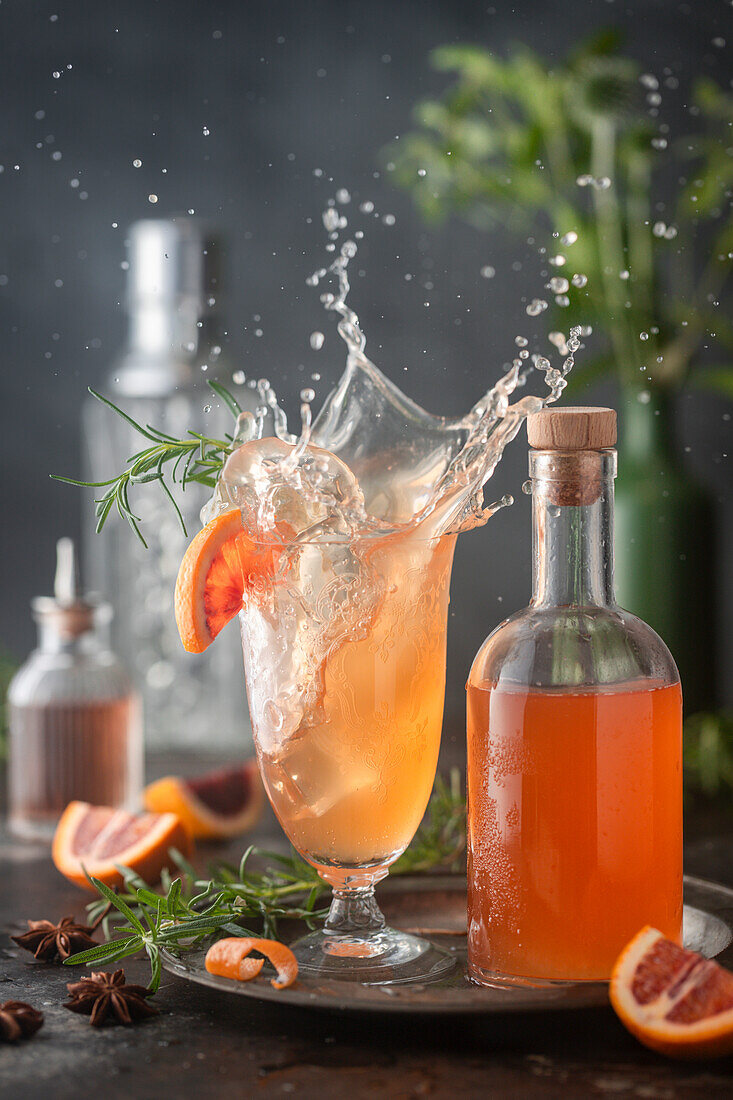 Action splash shot of ice falling into blood orange cocktail in vintage glass, with syrup bottle and citrus and rosemary garnish