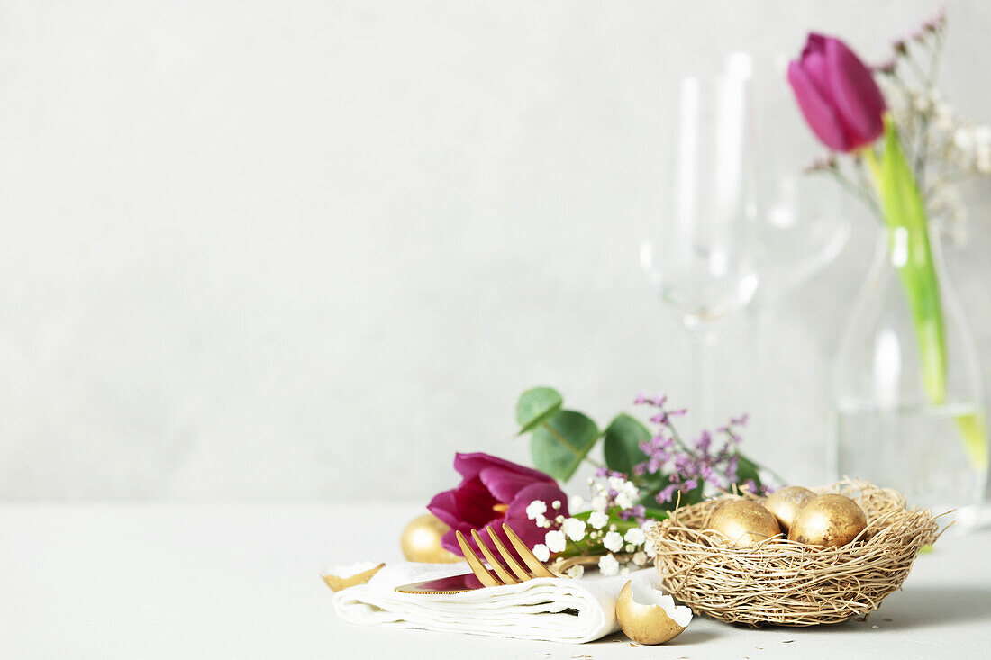 Ostertischdekoration mit Besteck, Frühlingsblumen und goldenen Eiern auf hellgrauem Hintergrund Kopierraum