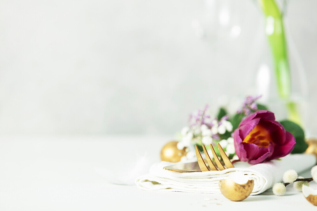 Easter table decorations with cutlery, spring flowers and golden eggs on light grey background copy space