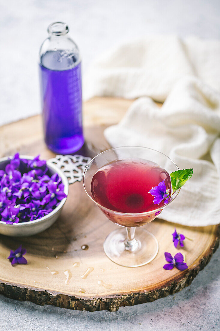 Pink cocktail in vintage glass with flower garnish, flowers and violet syrup on raw wood
