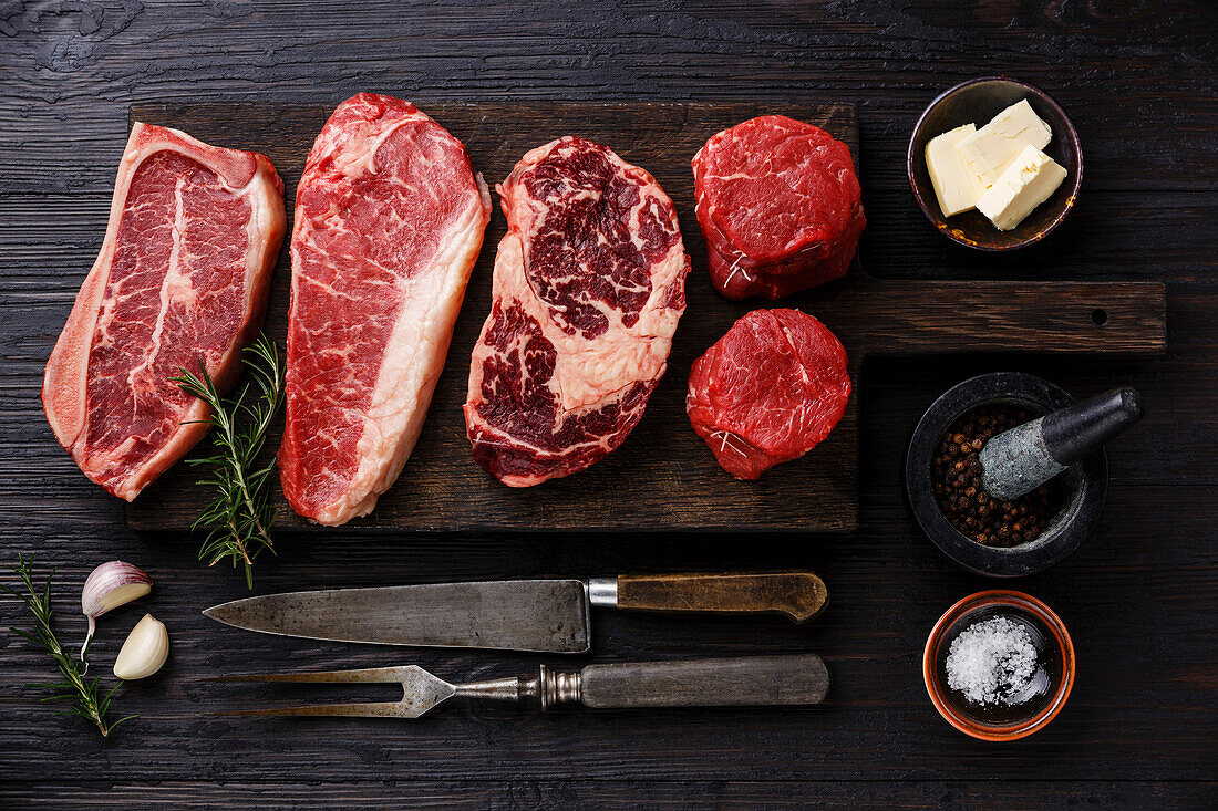 Various raw Black Angus prime steaks Blade on bone, striploin, rib eye, tenderloin filet mignon on wooden board and spices