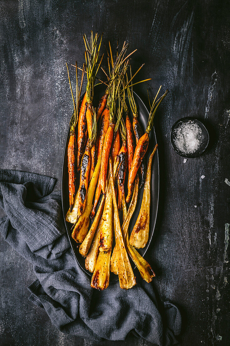 Roasted, glazed carrots and parsnips on a dark oval plate with grey linen napkin on a dark background